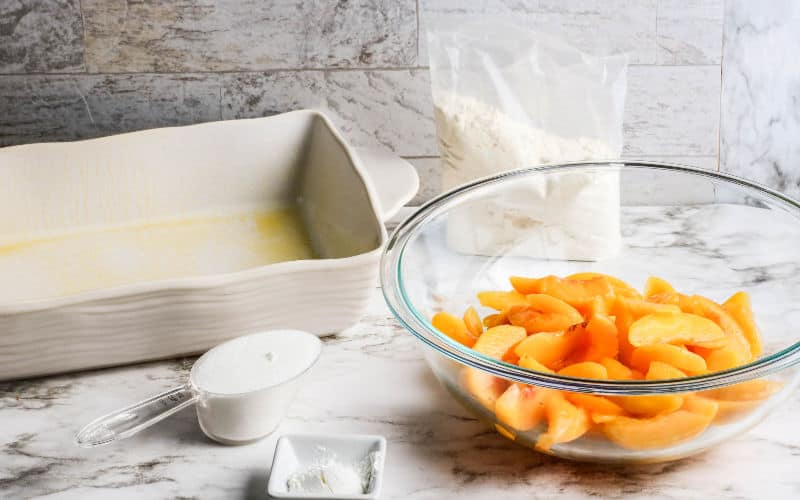 ingredients for a peach dump cake on table, peaches in bowl, casserole dish, sugar