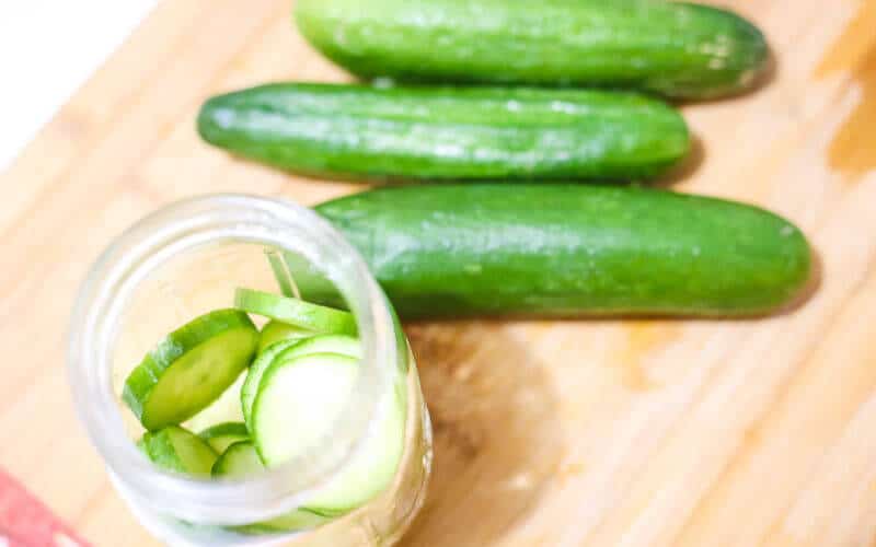 cucumbers chopped in coins in a ball jar on wooden cutting board with cucumbers ready to pickle